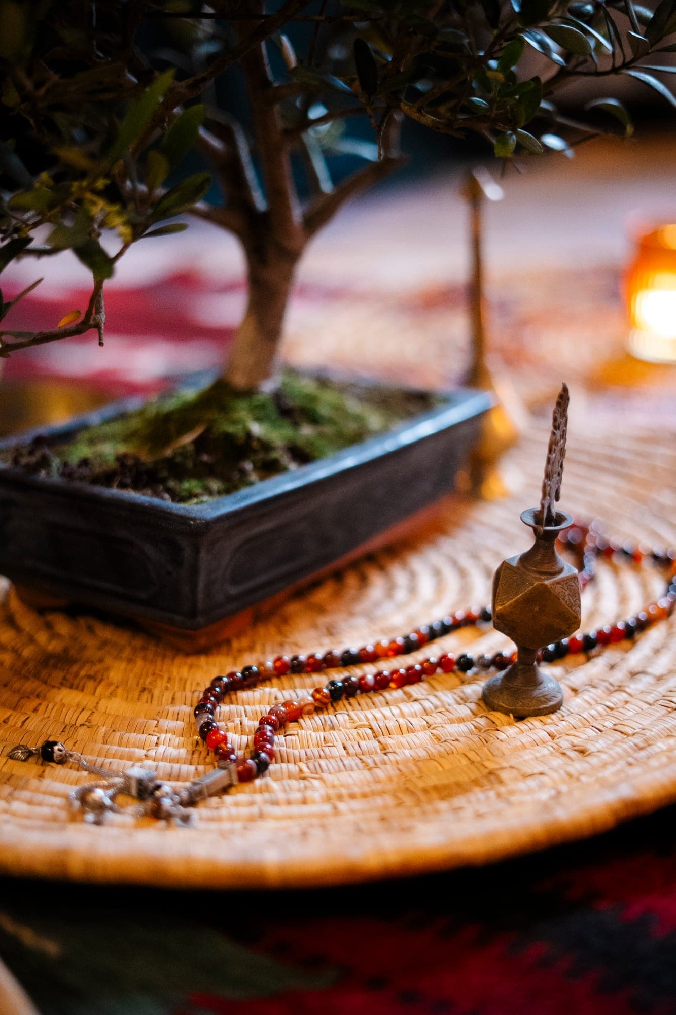 A bonsai and prayer beads on a woven table, a candle burning behind.