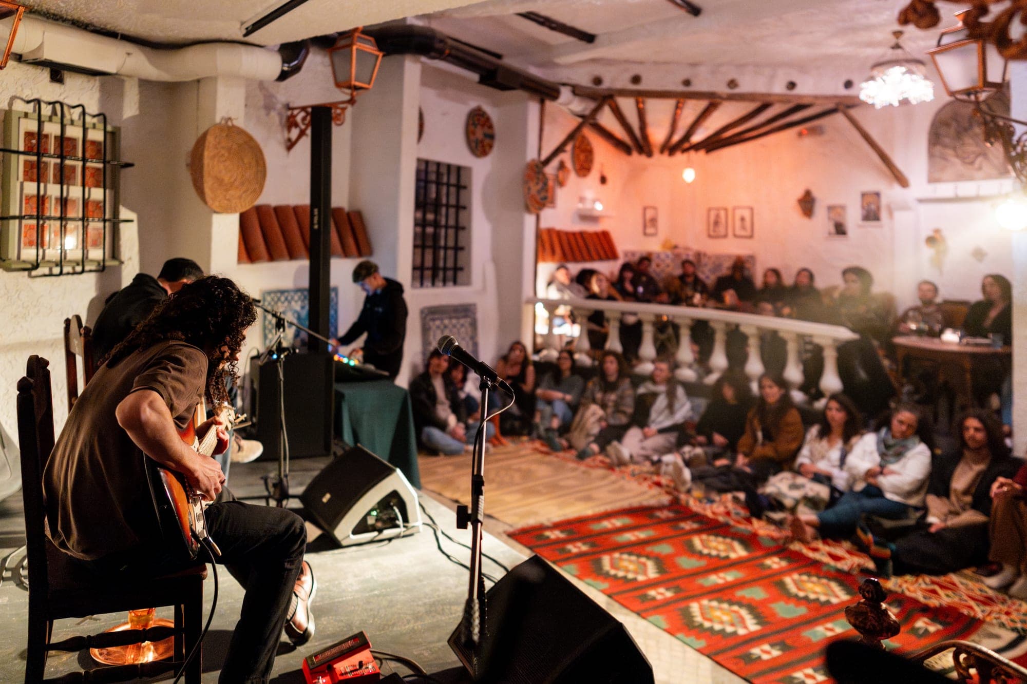 The same room by night — a musician on stage, the audience gathered on the rugs.
